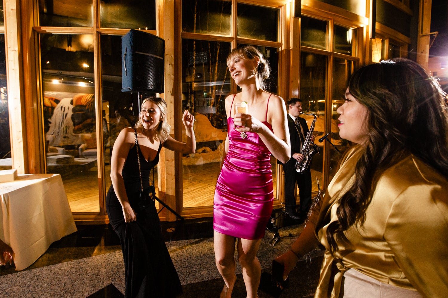 A lively party scene featuring women dancing and enjoying drinks in elegant attire, with a saxophonist performing in the background.