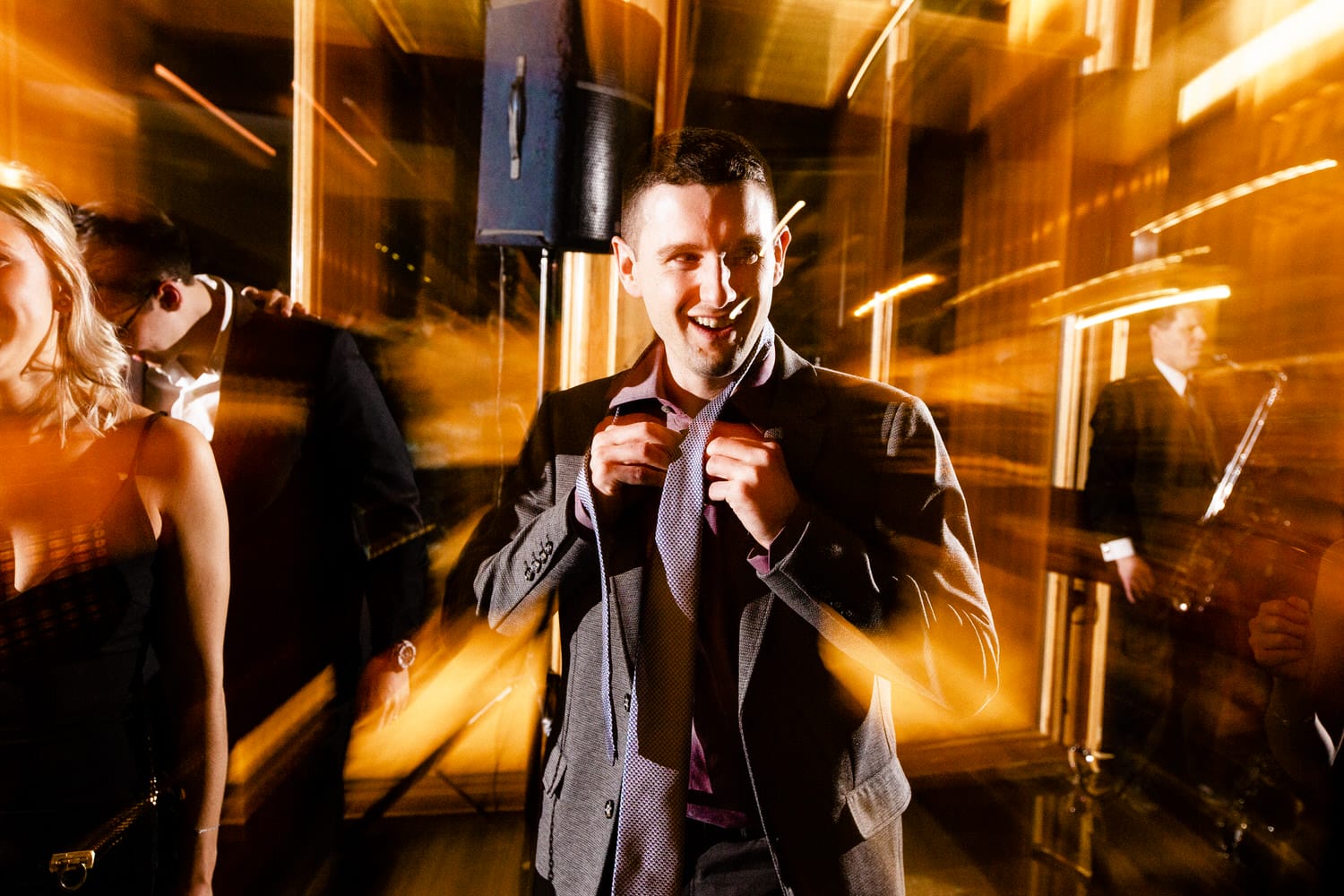 A man adjusting his tie and smiling, surrounded by a vibrant, lively atmosphere with light streaks suggesting movement and energy.