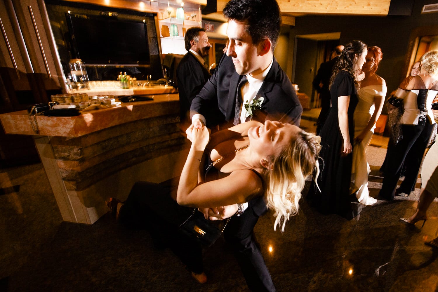 A man in a suit elegantly dips a woman in a black dress, both smiling joyfully amidst a festive atmosphere.