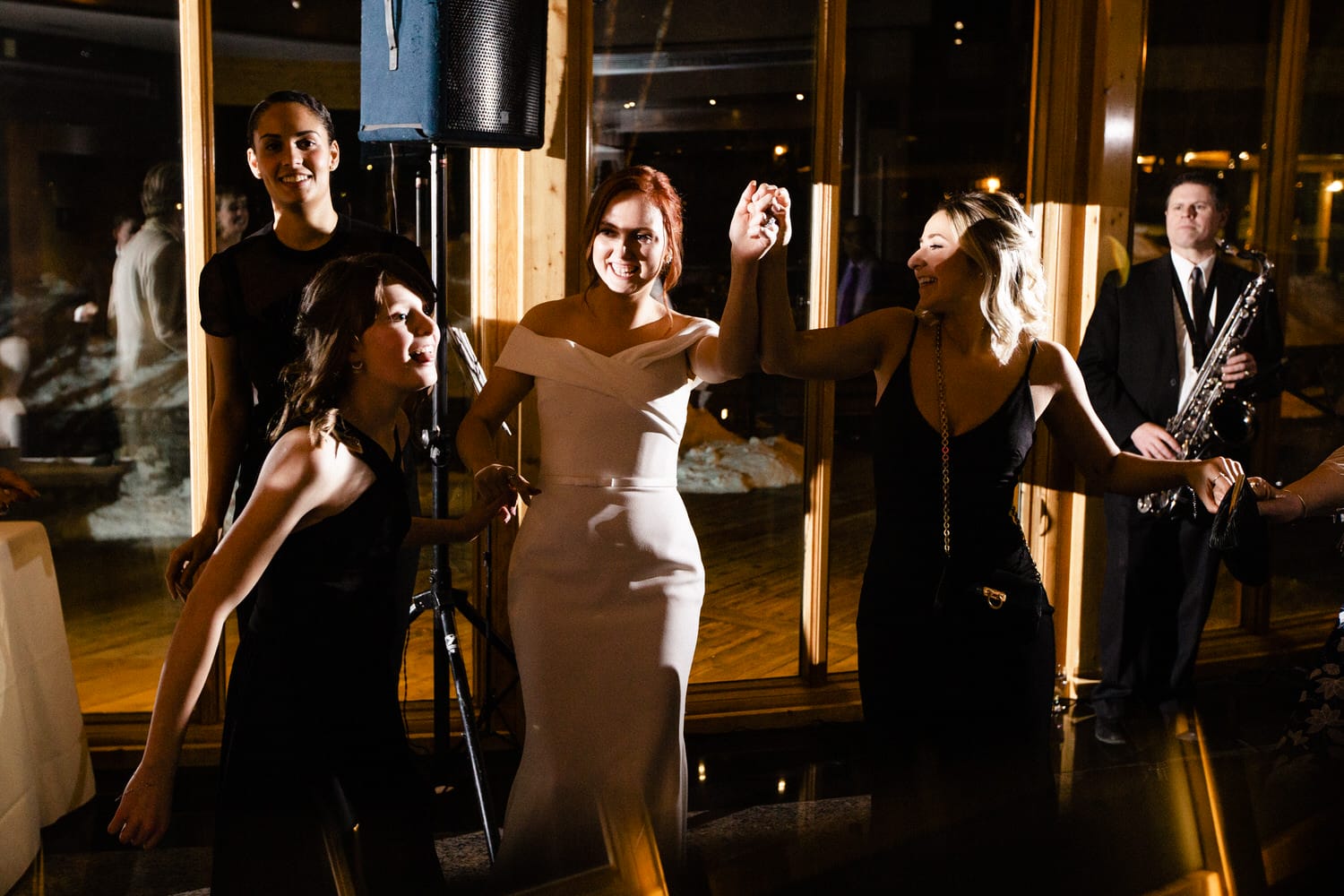 Group of women dancing joyfully at a festive event, with a live saxophonist in the background.