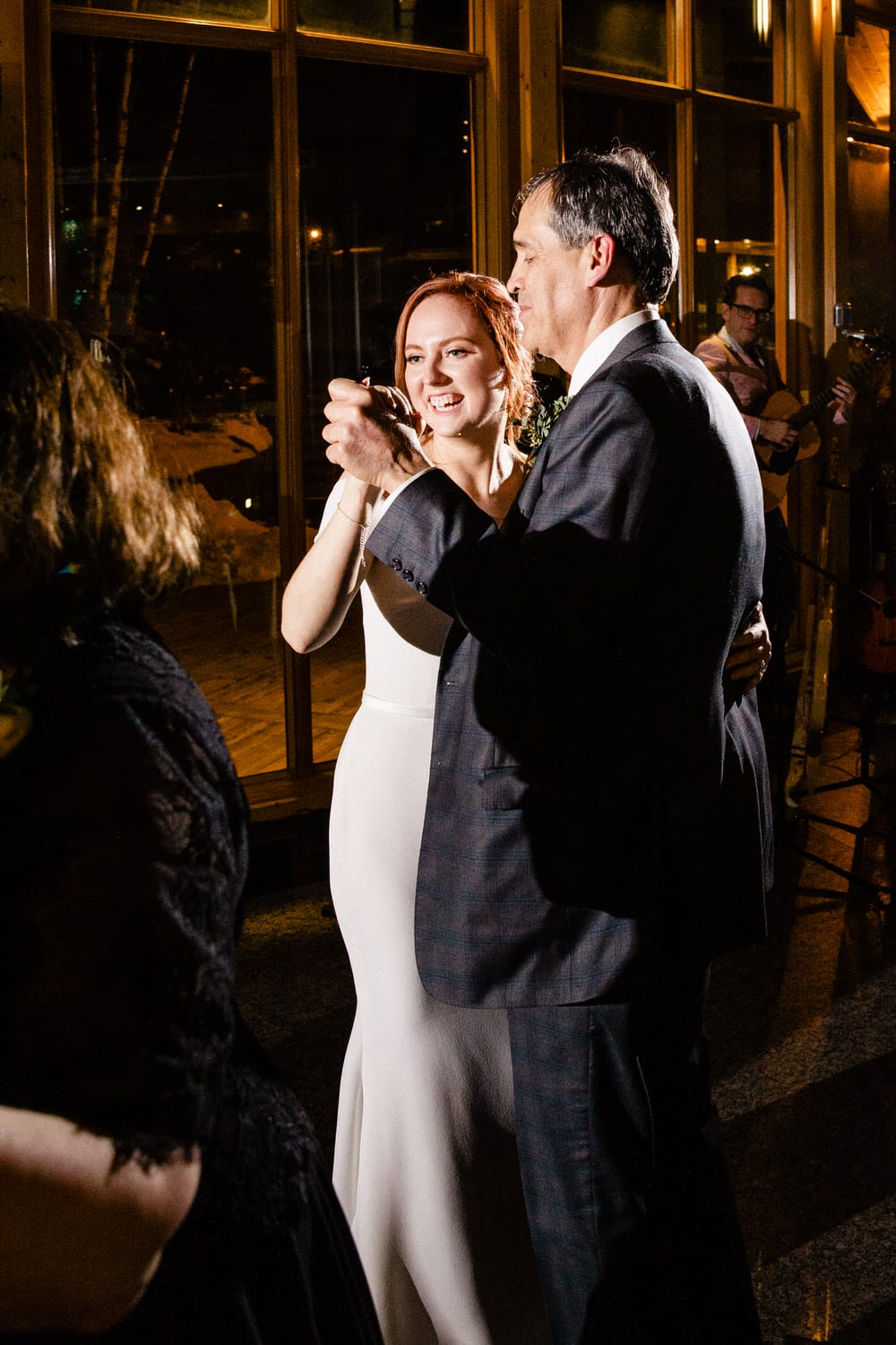 A bride and groom share a joyful dance together at their wedding reception, illuminated by warm lights and surrounded by guests.