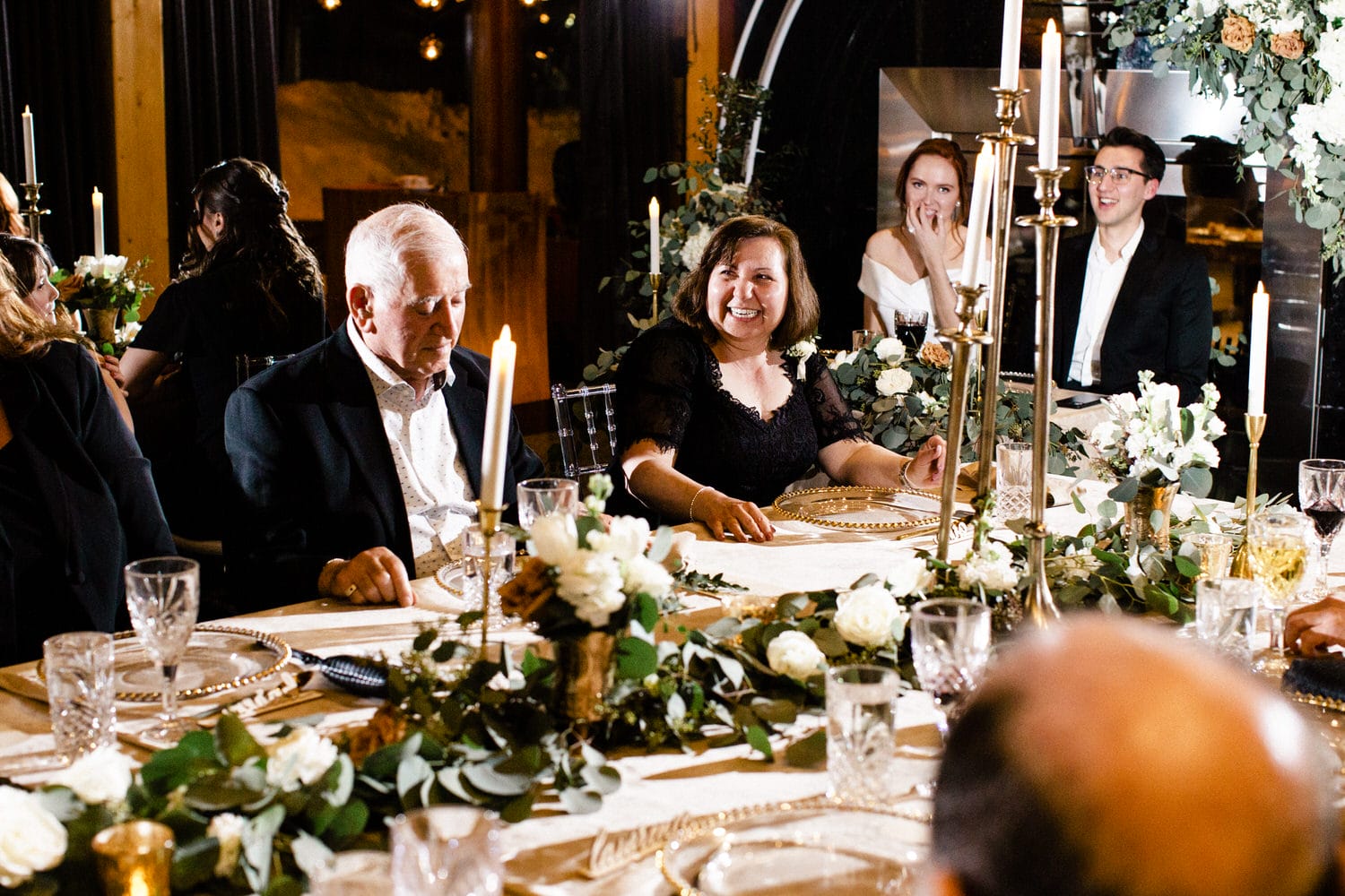 A joyful gathering around a beautifully set dinner table adorned with candles and floral arrangements, featuring smiling guests engaged in conversation.