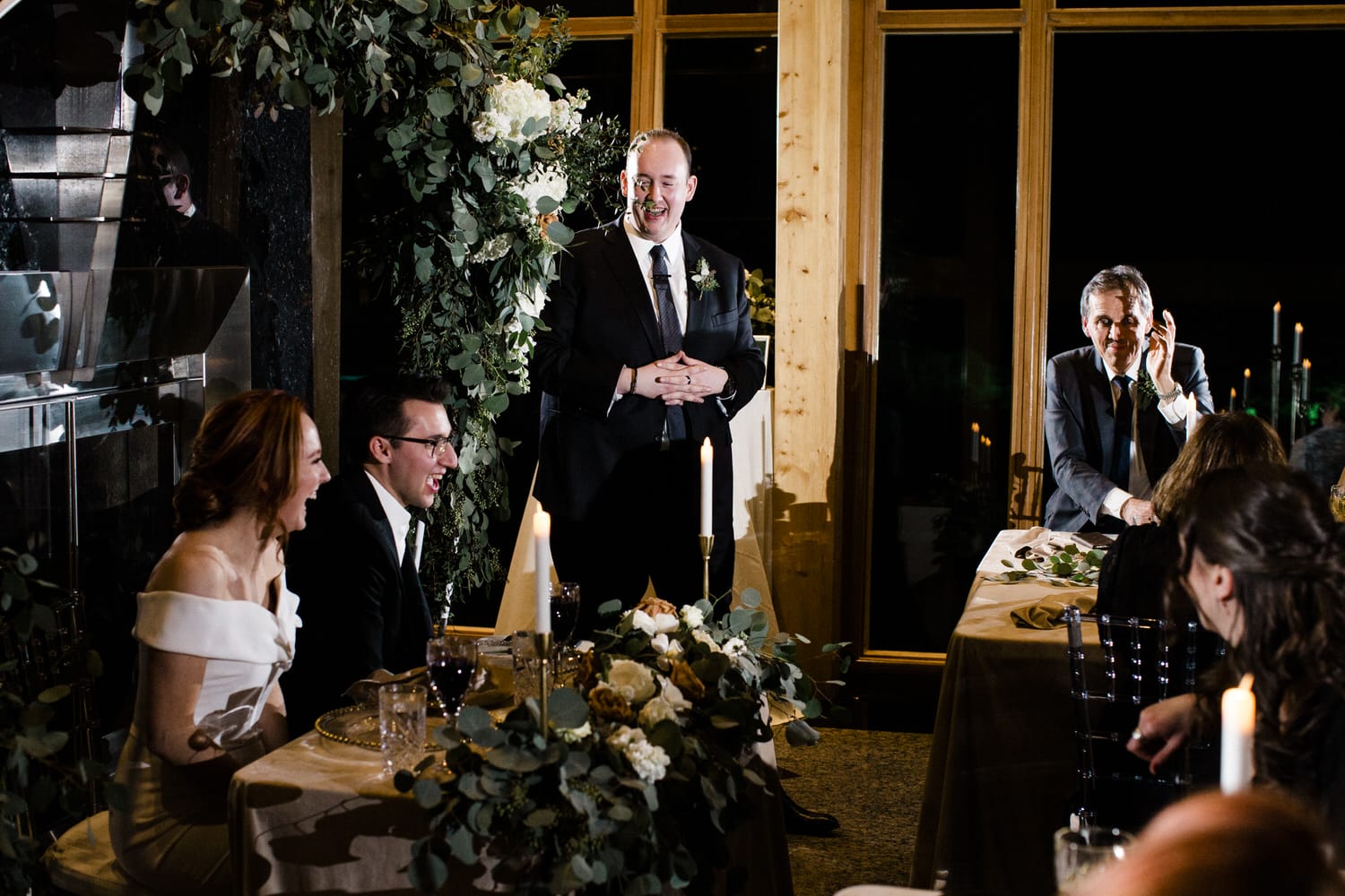 A joyful moment during a wedding reception, featuring a couple laughing while a speaker shares heartfelt words, surrounded by elegantly decorated tables and soft candlelight.