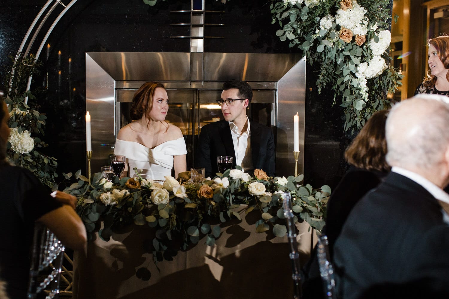 A bride and groom seated at a beautifully decorated table, surrounded by floral arrangements, with a glowing candle and an elegant fireplace in the background.