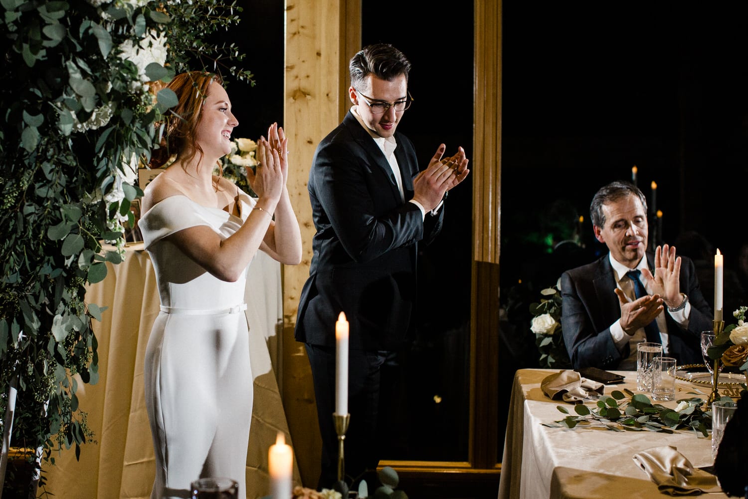 A bride and groom joyfully applaud during a wedding reception, surrounded by beautifully arranged flowers and candles.