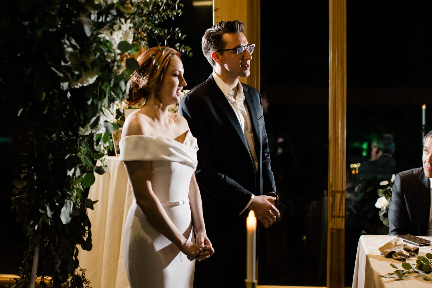 A couple stands together during their wedding ceremony, surrounded by elegant decor and a softly lit atmosphere.