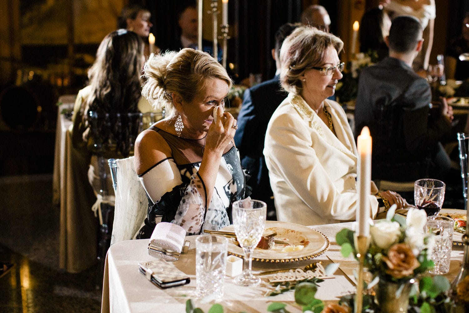 A woman wipes away tears during a heartfelt moment at a lavish event, seated beside another guest at a beautifully set table with candles and floral decor.