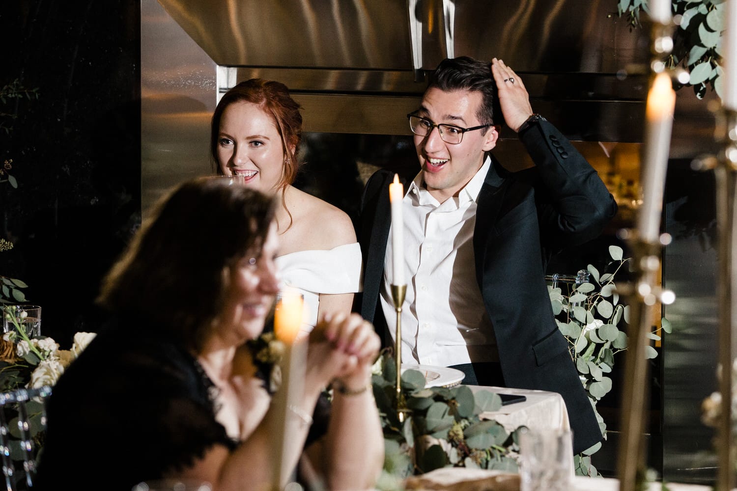 A couple laughing and enjoying a heartfelt moment during a wedding reception, with guests and candlelight in the foreground.