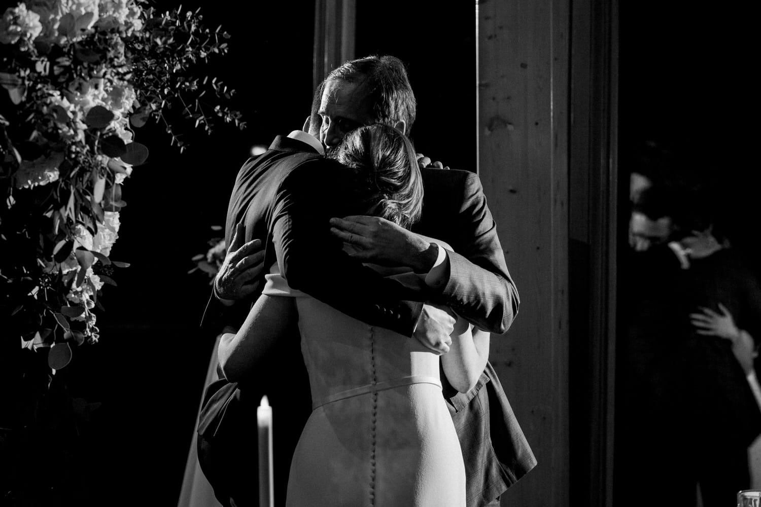 A man and a woman sharing a heartfelt hug in a softly lit setting, surrounded by floral arrangements and candlelight.