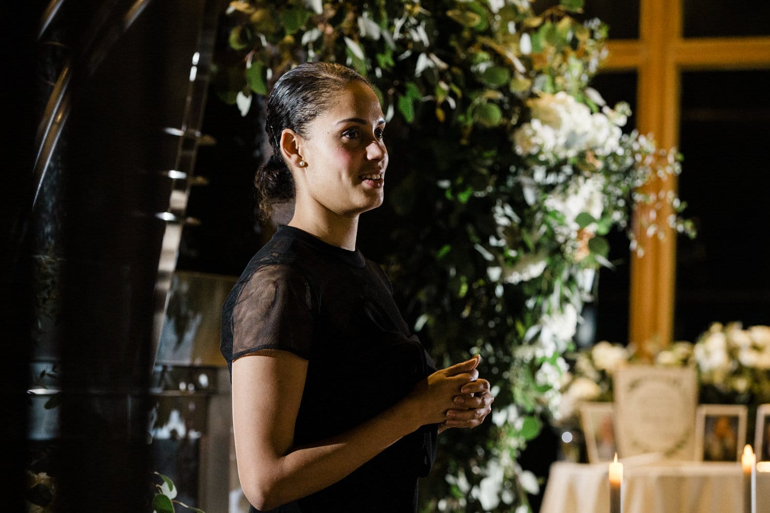 A woman in a black outfit speaks warmly in front of a floral backdrop during a celebration, with candles and decorations surrounding her.