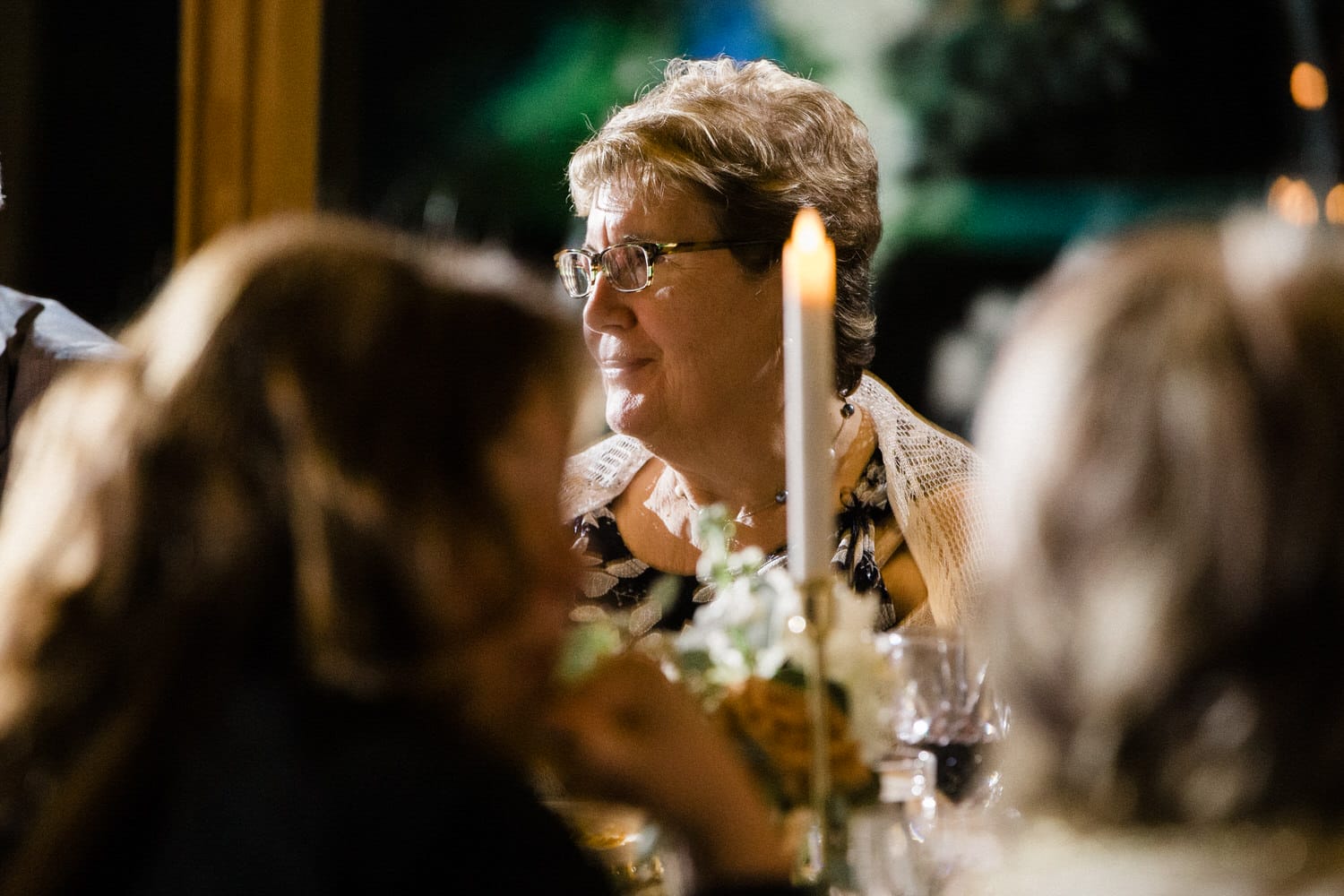 A woman with glasses enjoys a warm moment at a candlelit table amidst blurred company.