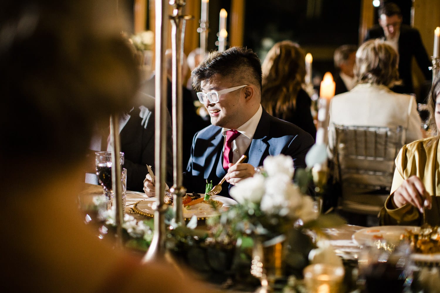 A man with glasses enjoys dinner at a beautifully arranged table with candles and elegant decor, surrounded by fellow diners.