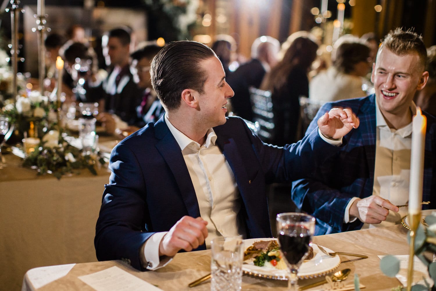 Two men engaging in conversation at a festive dinner table, with an elegant setting and guests in the background.