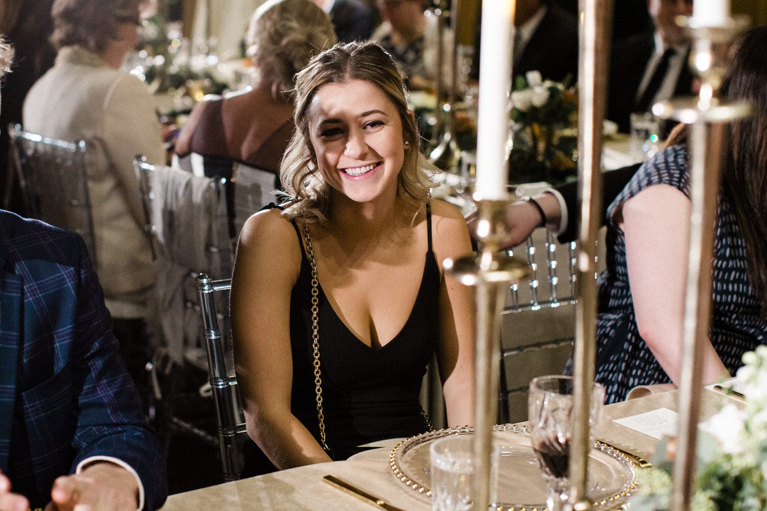 A smiling woman in a black dress sitting at a dining table during a festive event, surrounded by soft candlelight and guests in the background.