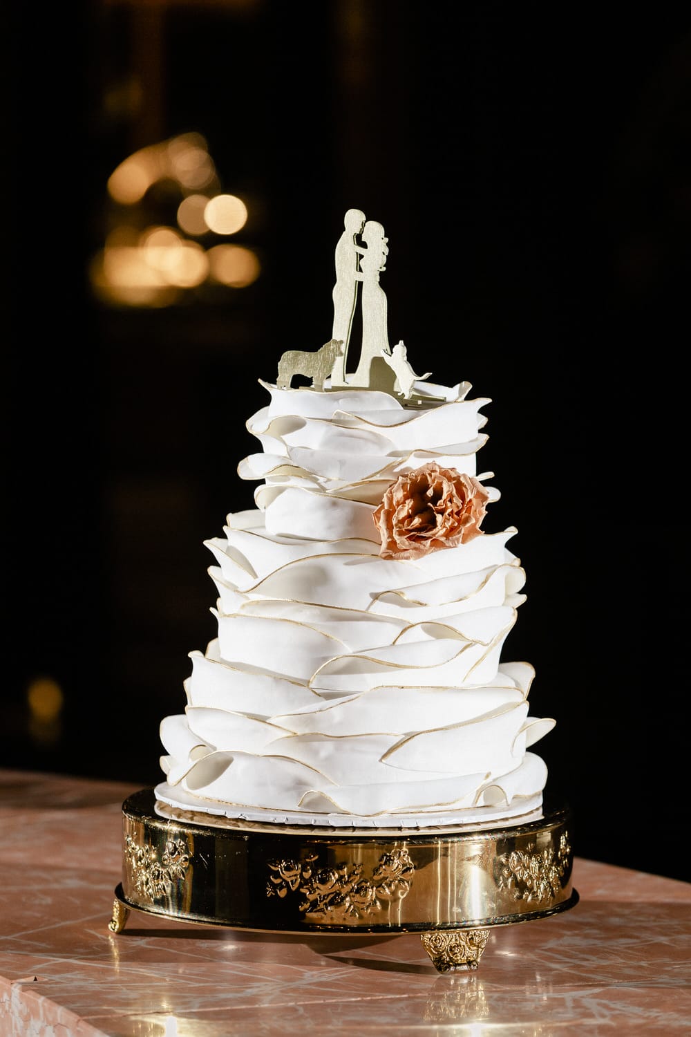 A multi-tiered wedding cake adorned with delicate ruffles and a golden cake topper depicting a couple and their pets, set on a decorative stand.