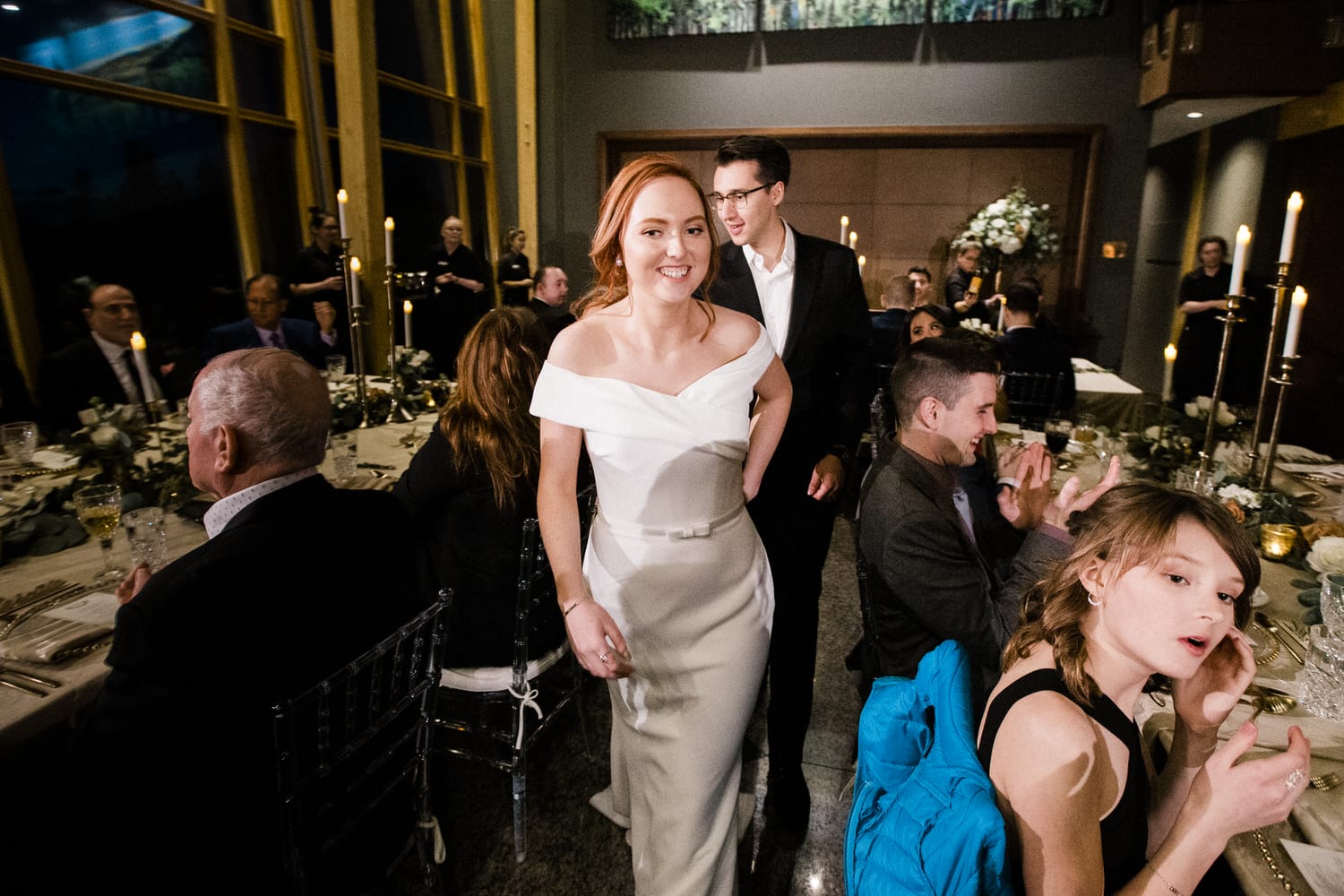 A smiling woman in a white dress walks through a beautifully arranged dining area filled with guests, candles, and floral decorations.