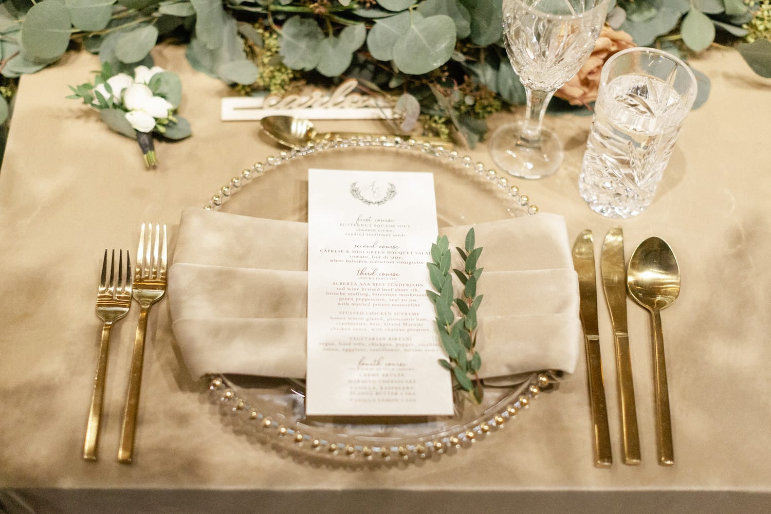 A beautifully arranged dinner setting featuring a glass plate, gold cutlery, a menu, and decorative greenery on a beige tablecloth.