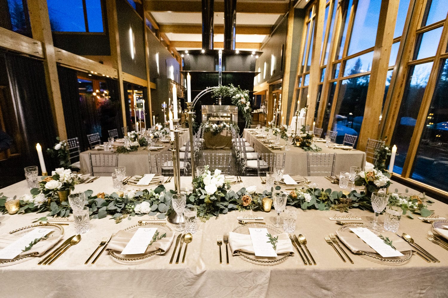 A beautifully arranged dining table featuring floral centerpieces, candles, and elegant tableware in a modern setting with large windows and warm lighting.