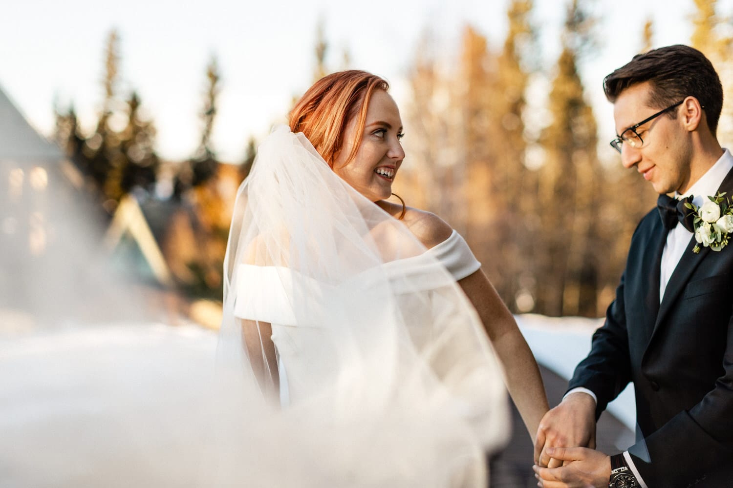 Wedding Bliss in Nature::A bride and groom share a joyful moment outdoors, surrounded by trees and soft sunlight, with the bride's veil flowing elegantly in the breeze.