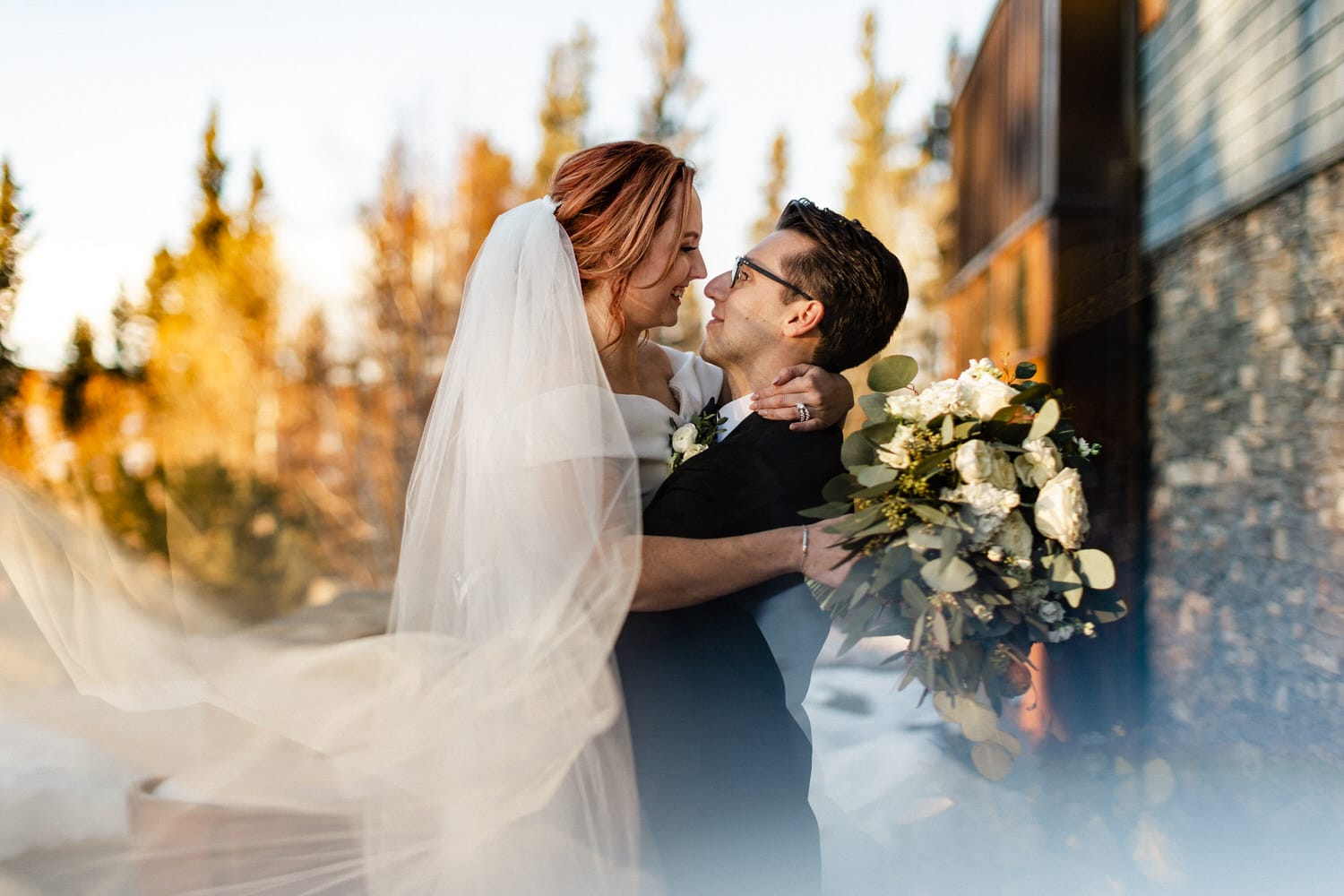 A couple shares a tender moment outdoors, surrounded by a picturesque autumn landscape, with the bride holding a bouquet of white flowers.