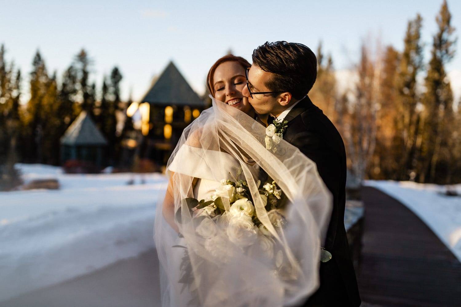 A joyful couple embracing outdoors, with the bride’s veil flowing softly in the breeze, surrounded by snow and trees in the background.