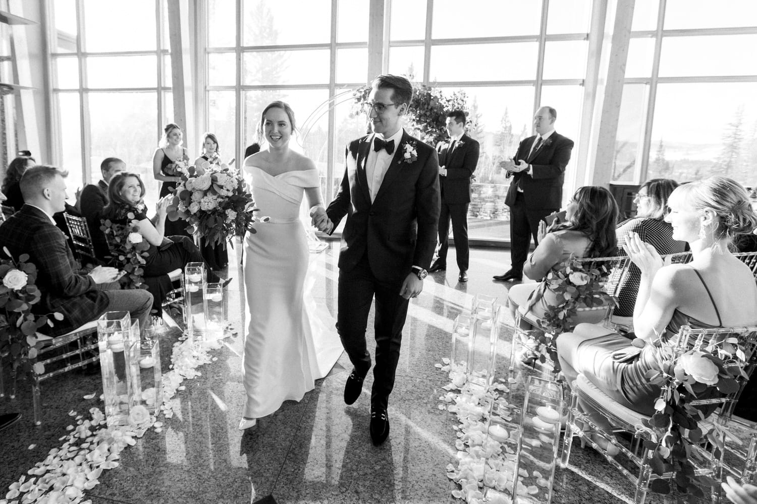 A happy couple walks down the aisle during their wedding ceremony, surrounded by applauding guests in a beautifully decorated venue.