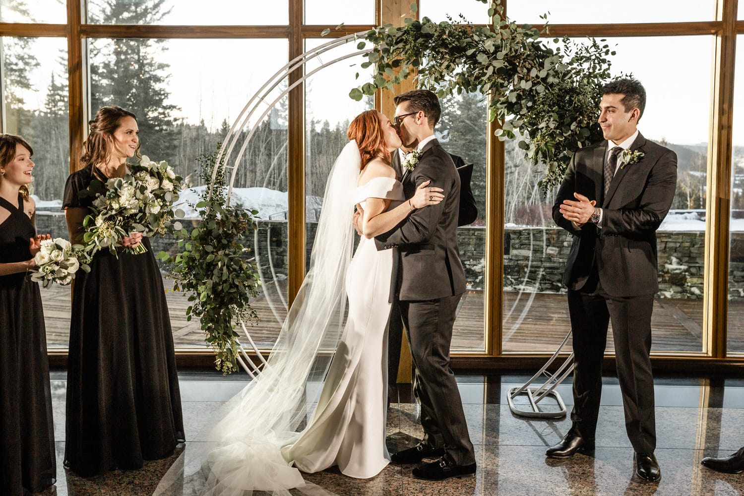 A couple shares a tender kiss during their wedding ceremony, surrounded by bridesmaids and a lush floral arch.