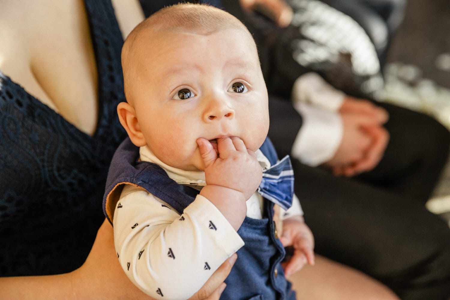 A close-up of a baby wearing a bow tie and grabbing his fingers, looking curiously at the camera.