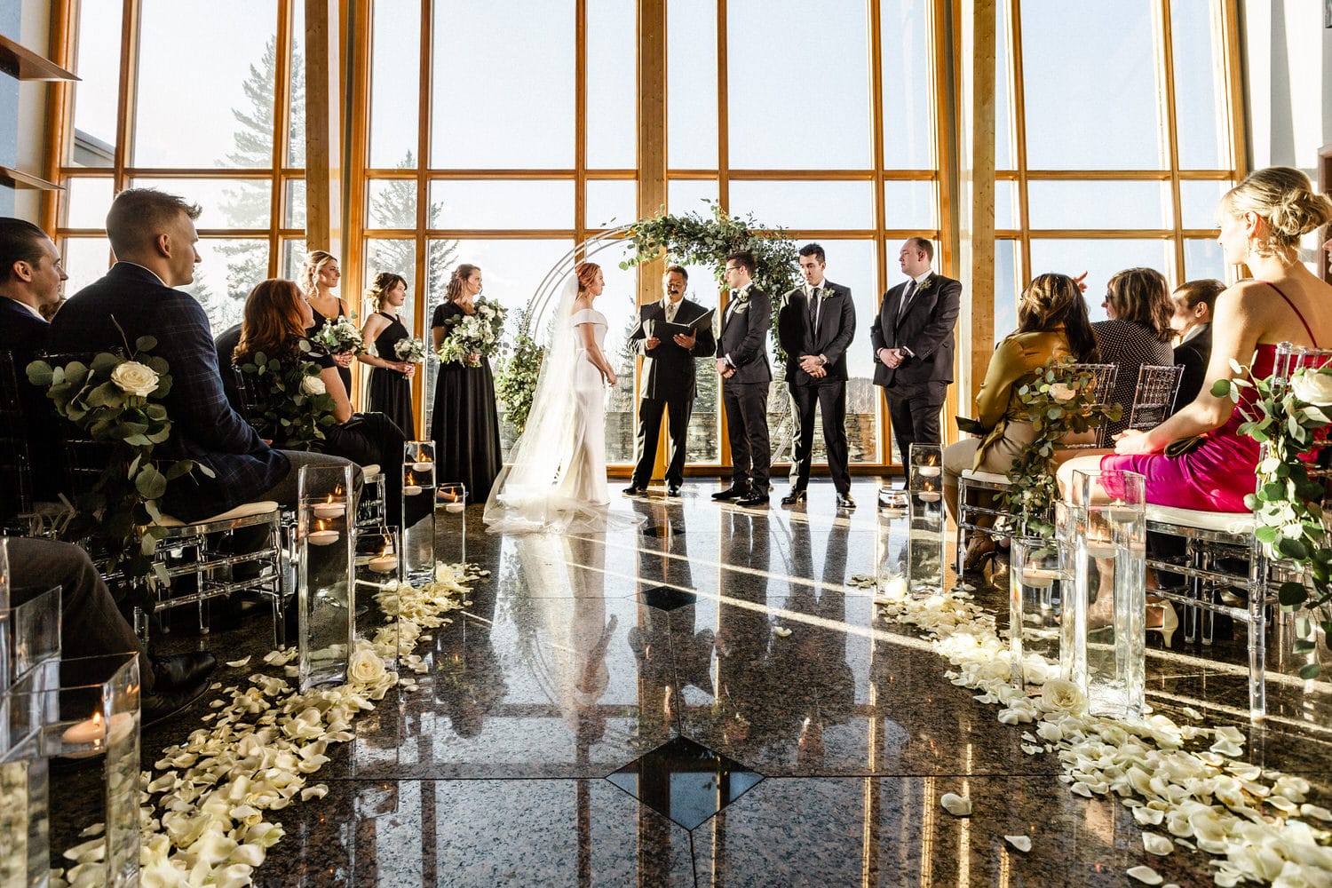 A bride and groom exchange vows surrounded by guests and wedding party in a beautifully decorated hall with natural light.