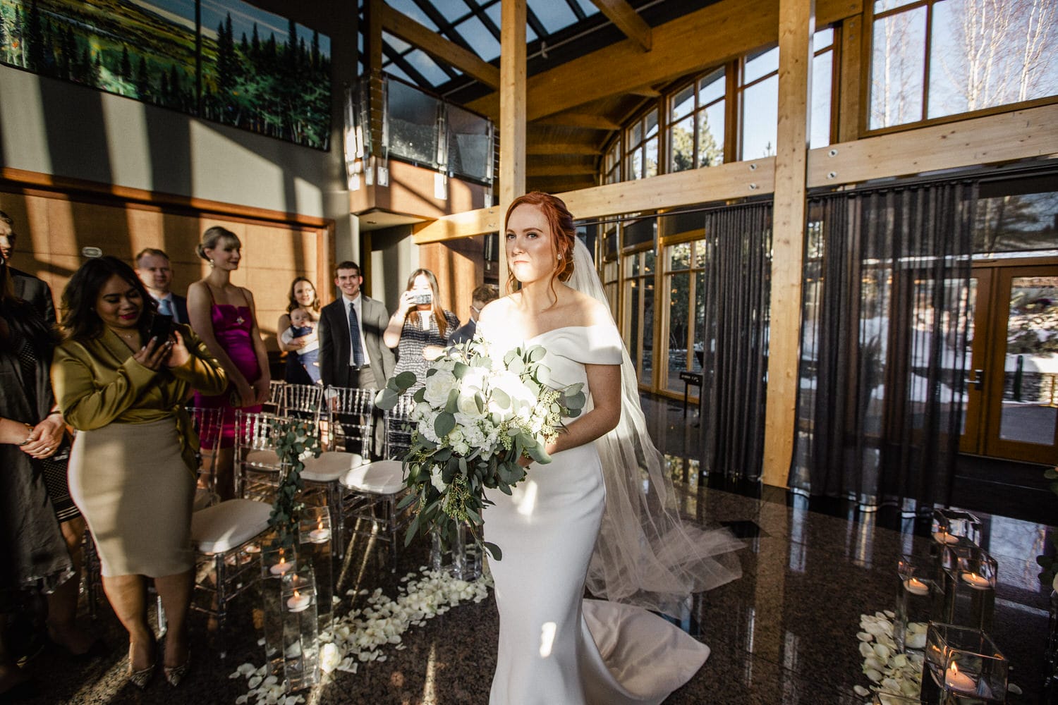 A bride in a sleek white gown and veil walks down the aisle, holding a bouquet, as guests look on with interest in a beautifully lit venue.