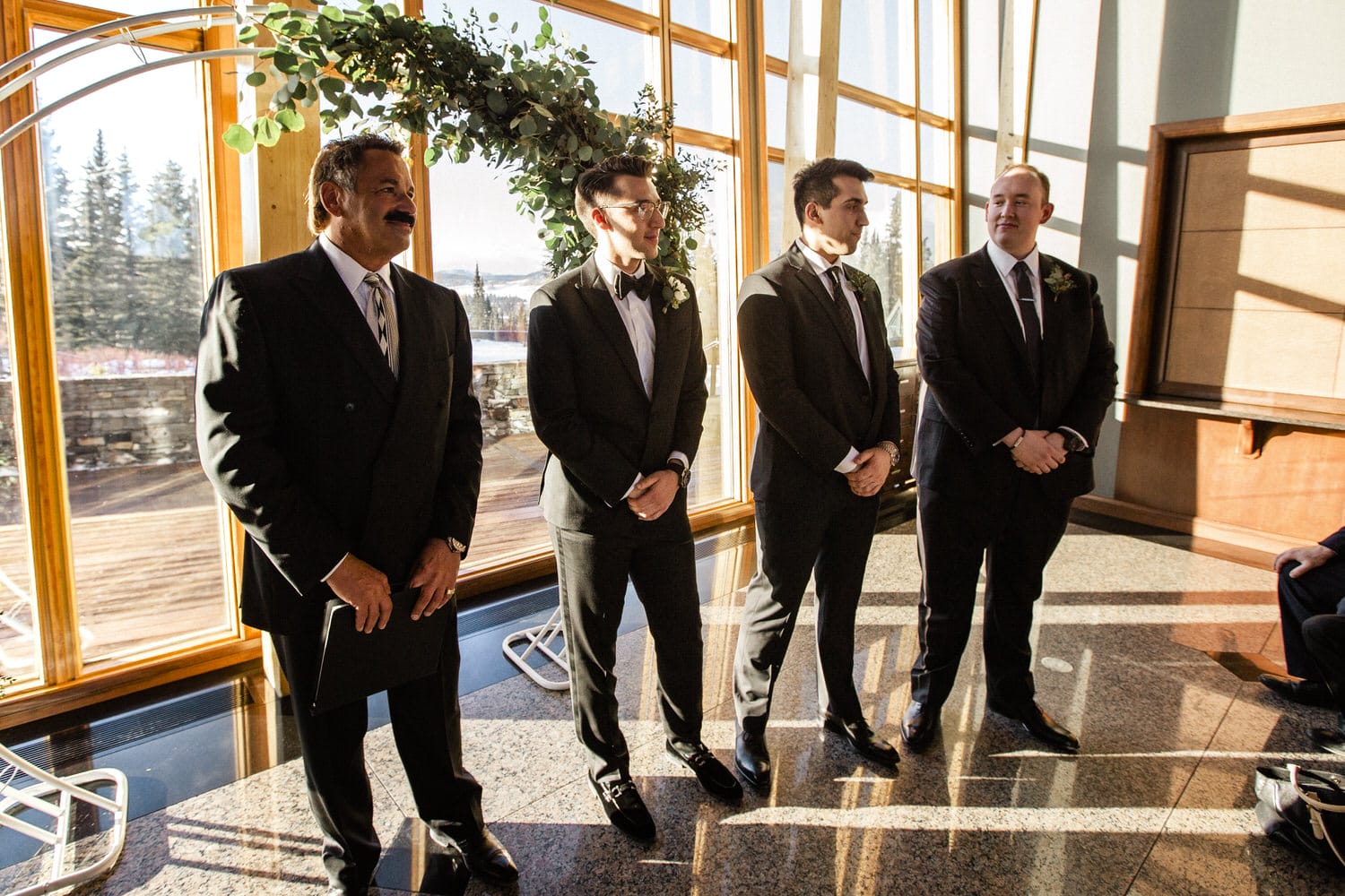 Four men in formal attire stand in a bright, elegant space with a wooden arch and greenery, preparing for a wedding ceremony.