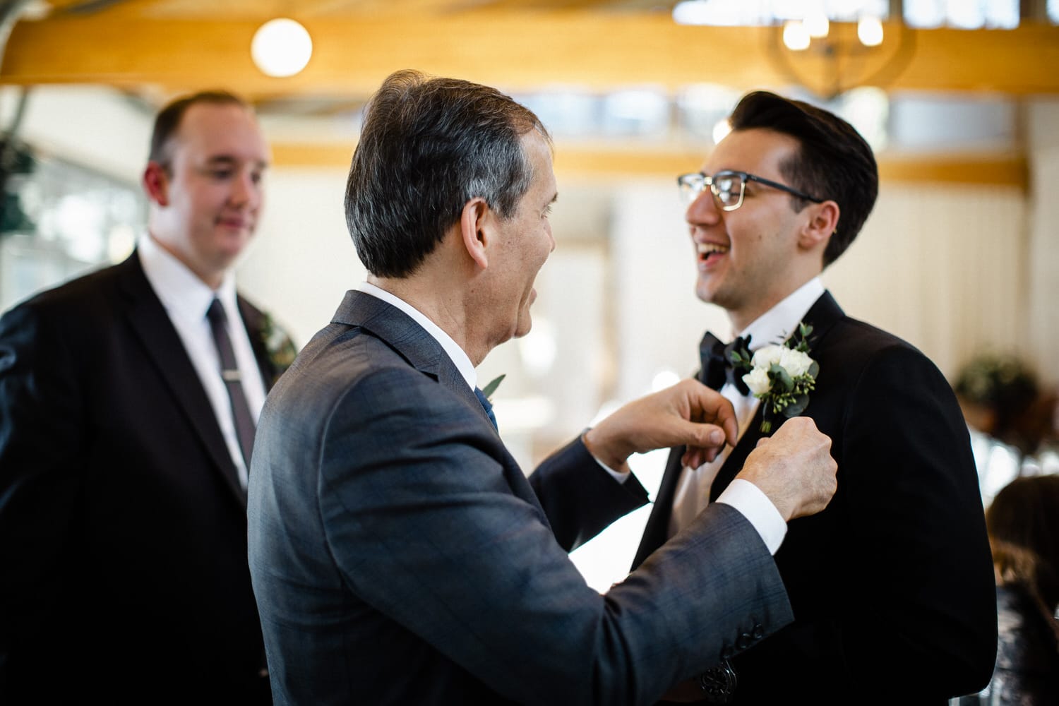 A groom and his father share a joyful moment as they adjust flower lapels before the wedding ceremony, with a third man in the background.