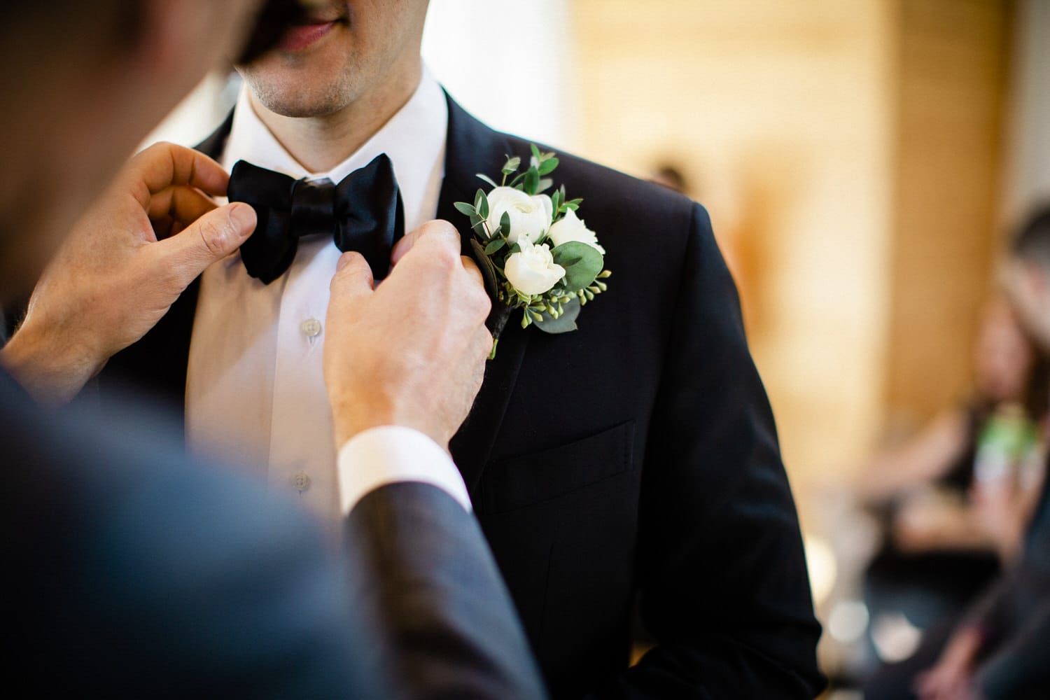 A man adjusts a bow tie on another man's formal attire, showcasing a boutonniere on the lapel in a celebratory setting.