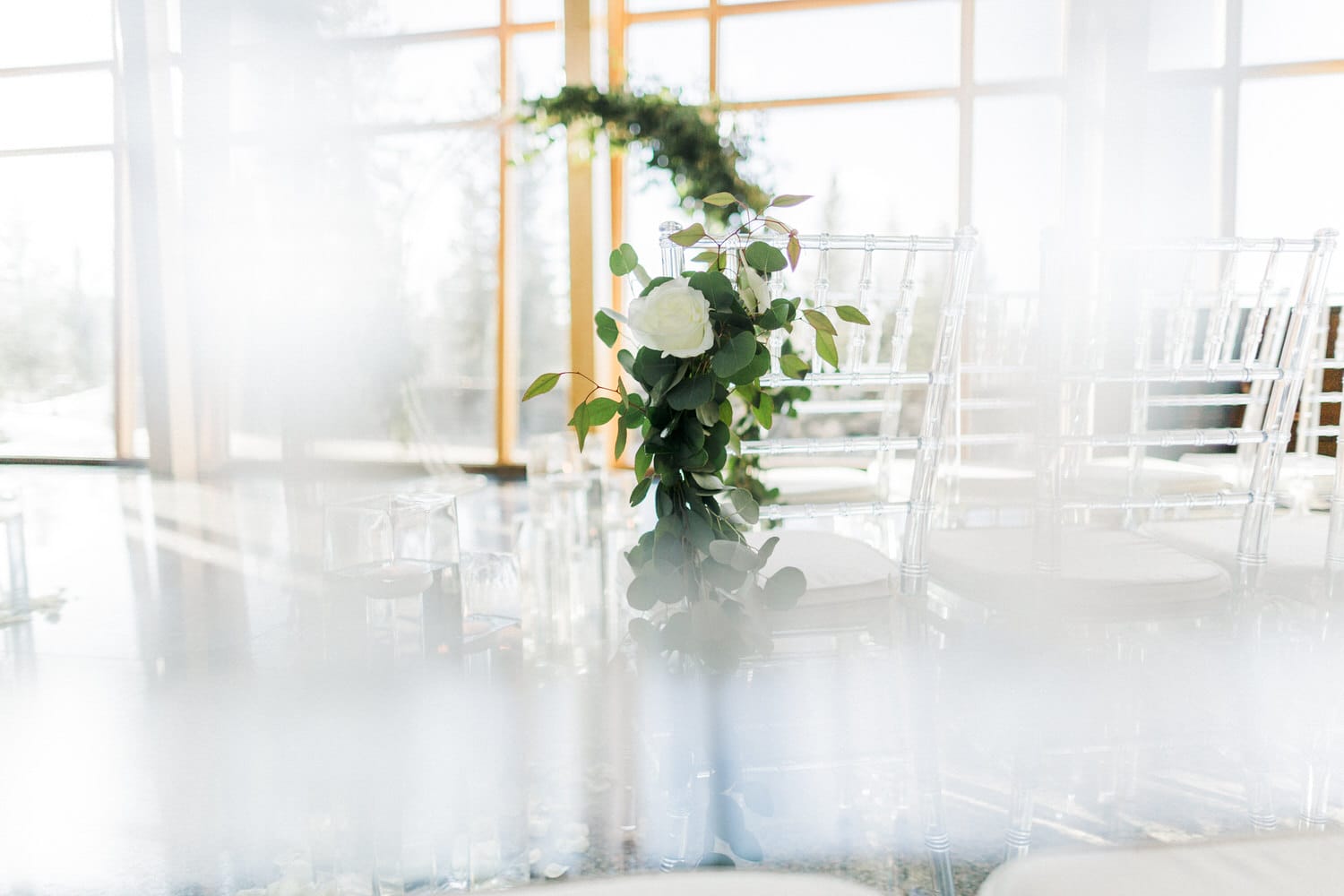 A close-up view of a beautifully arranged floral centerpiece with greenery and a white rose, set against a backdrop of stylish transparent chairs and large windows letting in natural light.