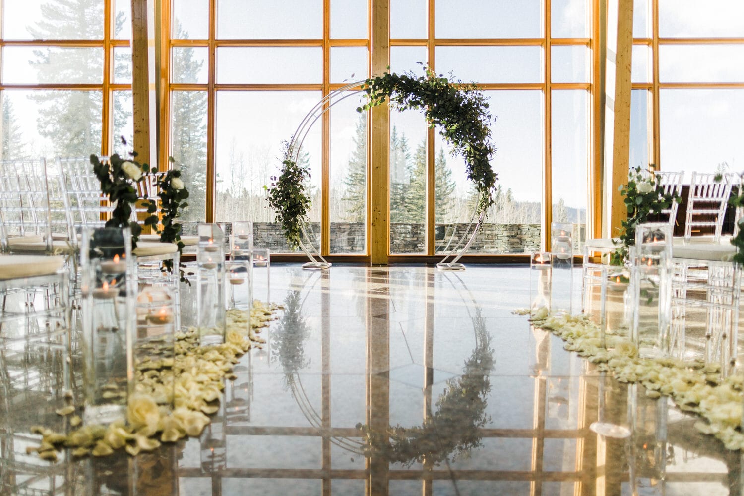 A beautifully arranged wedding ceremony space featuring transparent chairs, floral decorations, and a lush archway, set against large windows with a scenic view.