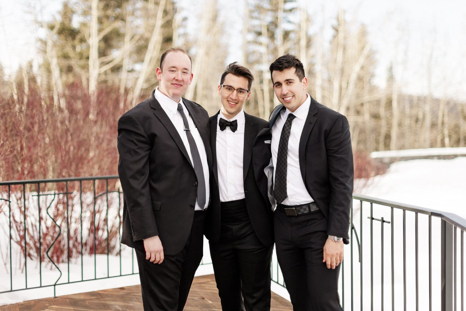 Three well-dressed men in black suits pose together outdoors with a snowy landscape and trees in the background.