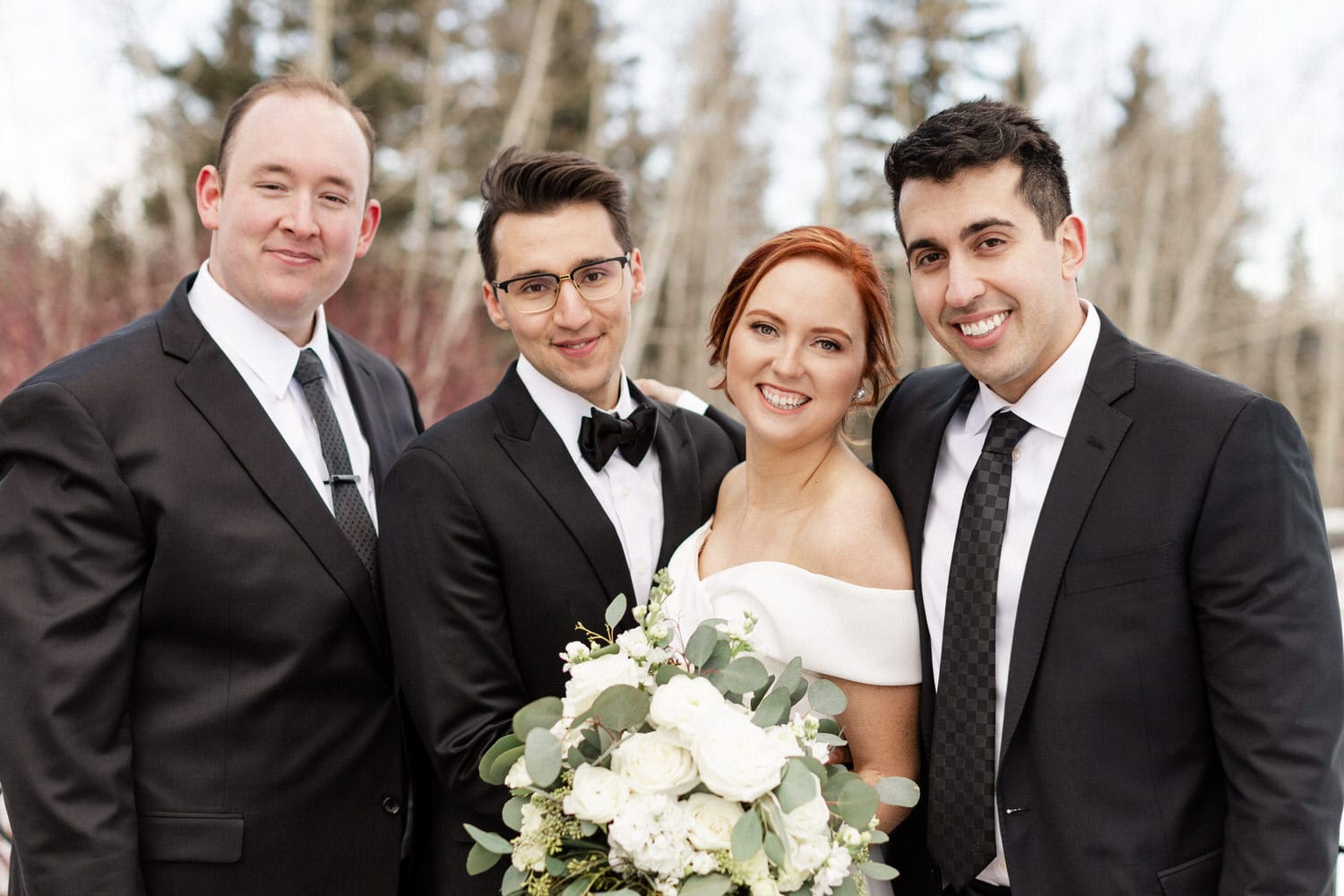 A joyful couple poses with friends, showcasing a beautiful bouquet and elegant wedding attire against a serene outdoor backdrop.