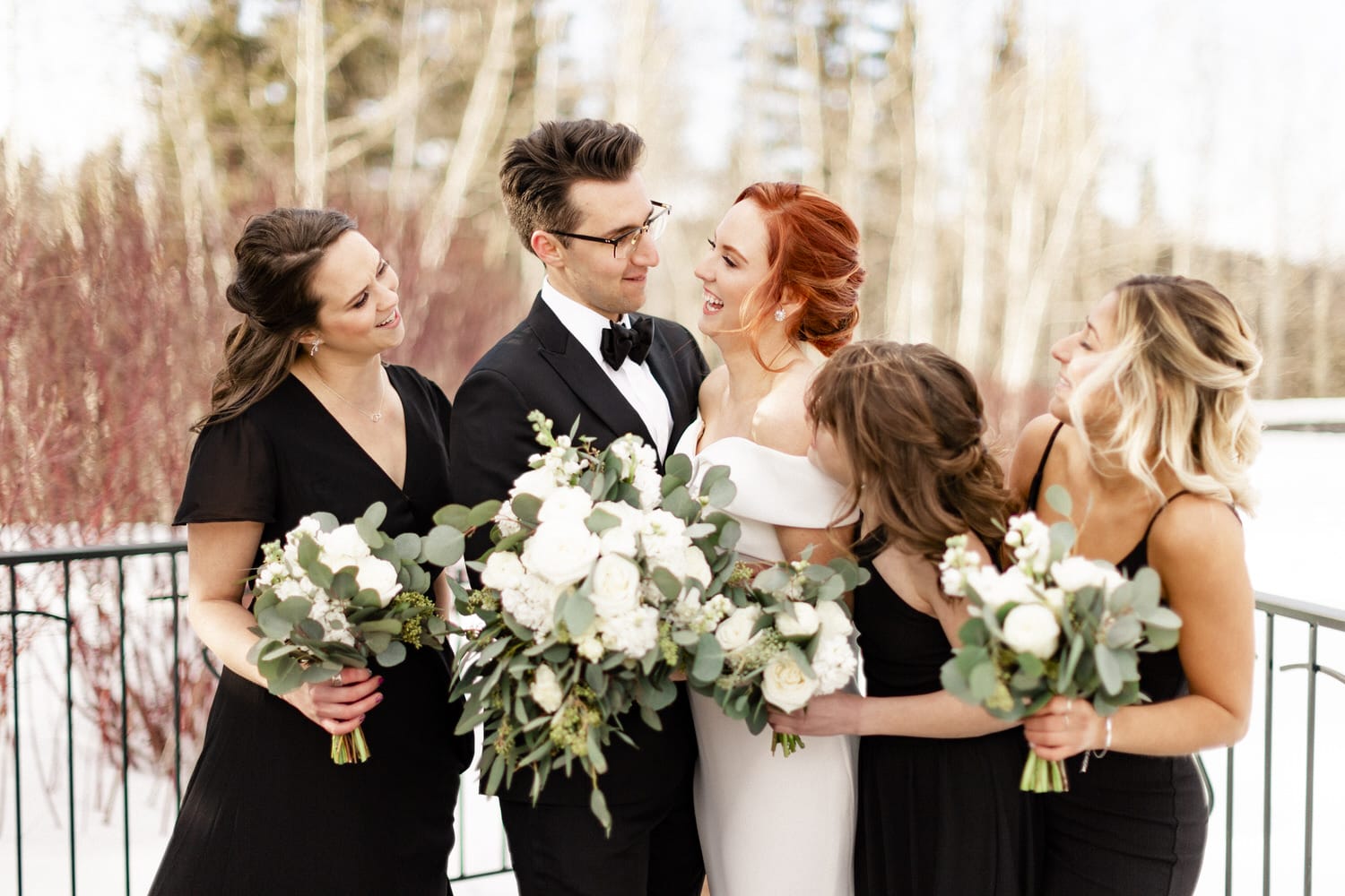 A bride in a white dress and her bridesmaids in black gowns, all laughing and holding white floral bouquets, surrounded by a snowy landscape.