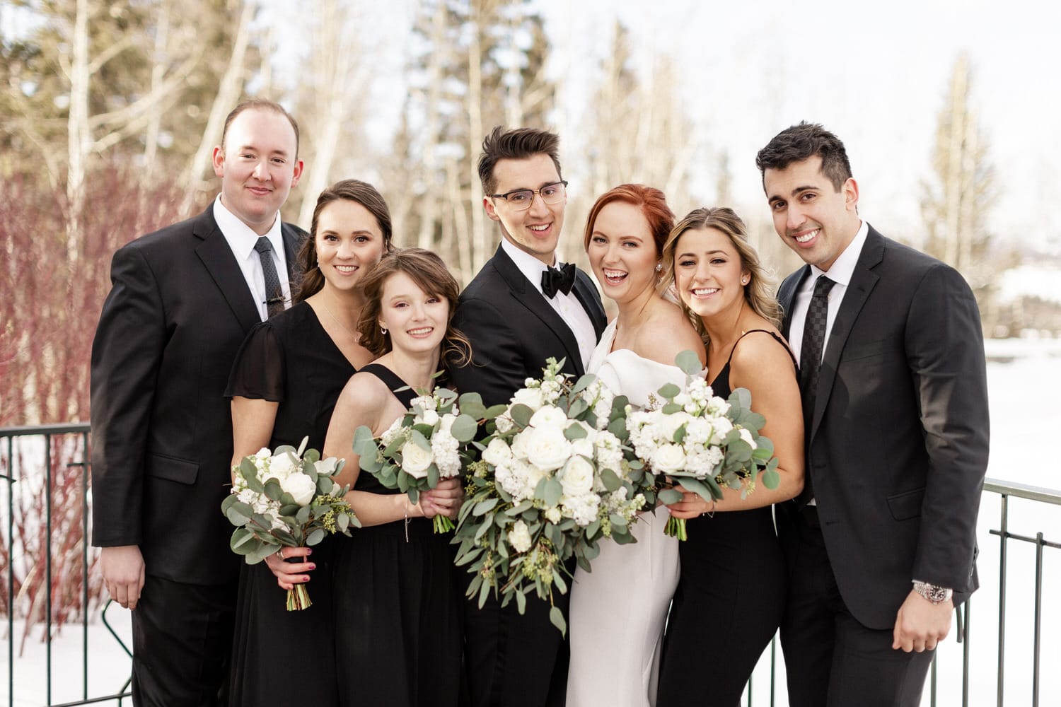 A joyful wedding party poses together outdoors, holding bouquets of white flowers and greenery, dressed in formal black attire.