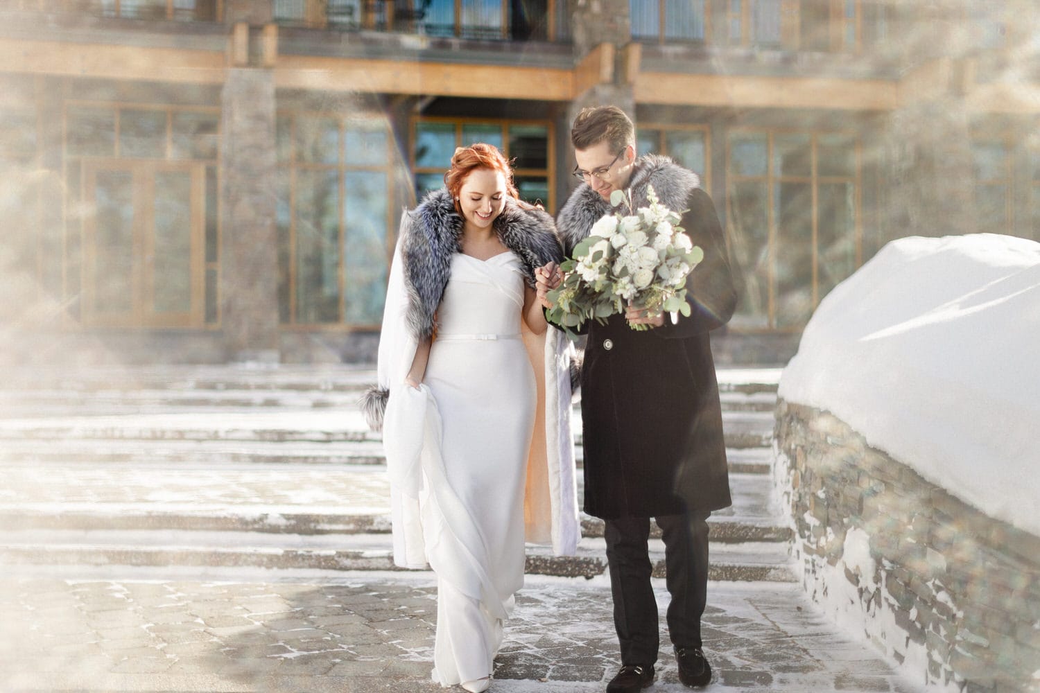 A cheerful couple walking hand in hand on a snowy staircase, with the bride in a stunning white dress and elegant fur shawl, while the groom holds a bouquet of flowers.