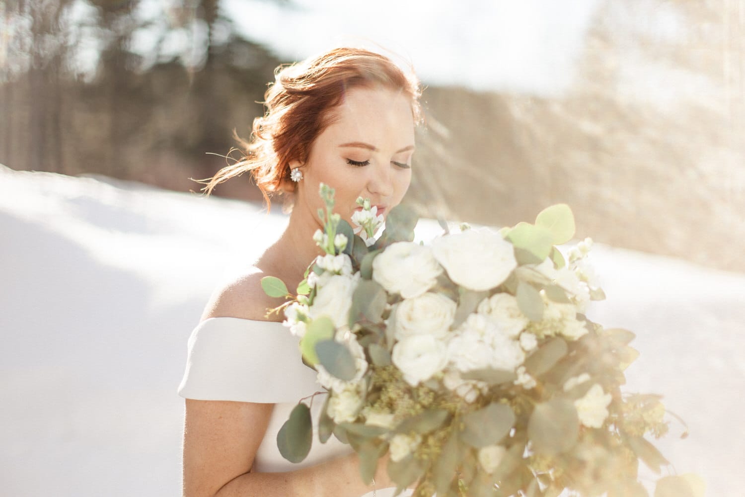 A bride with red hair softly smiles as she holds a lush bouquet of white flowers, surrounded by a snowy landscape in soft sunlight.