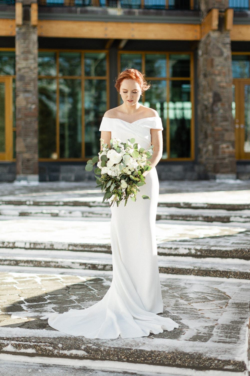 A bride in a flowing white gown stands on stone steps, holding a lush bouquet of white flowers and greenery, with a serene expression.