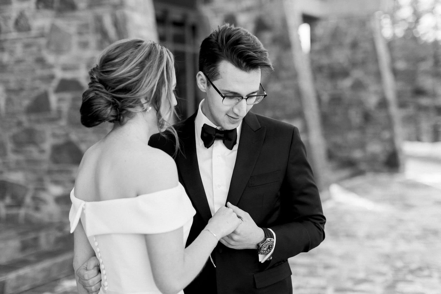 A groom in a tuxedo gazes at his bride, who is wearing an off-the-shoulder wedding dress, as they hold hands in a romantic setting.