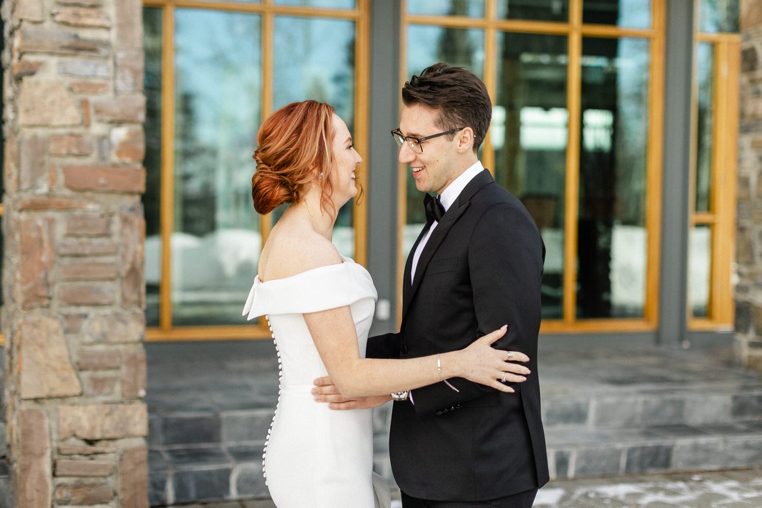 A joyful bride in an off-shoulder white dress and a groom in a black tuxedo share a loving moment outdoors, with a stone building and large windows in the background.