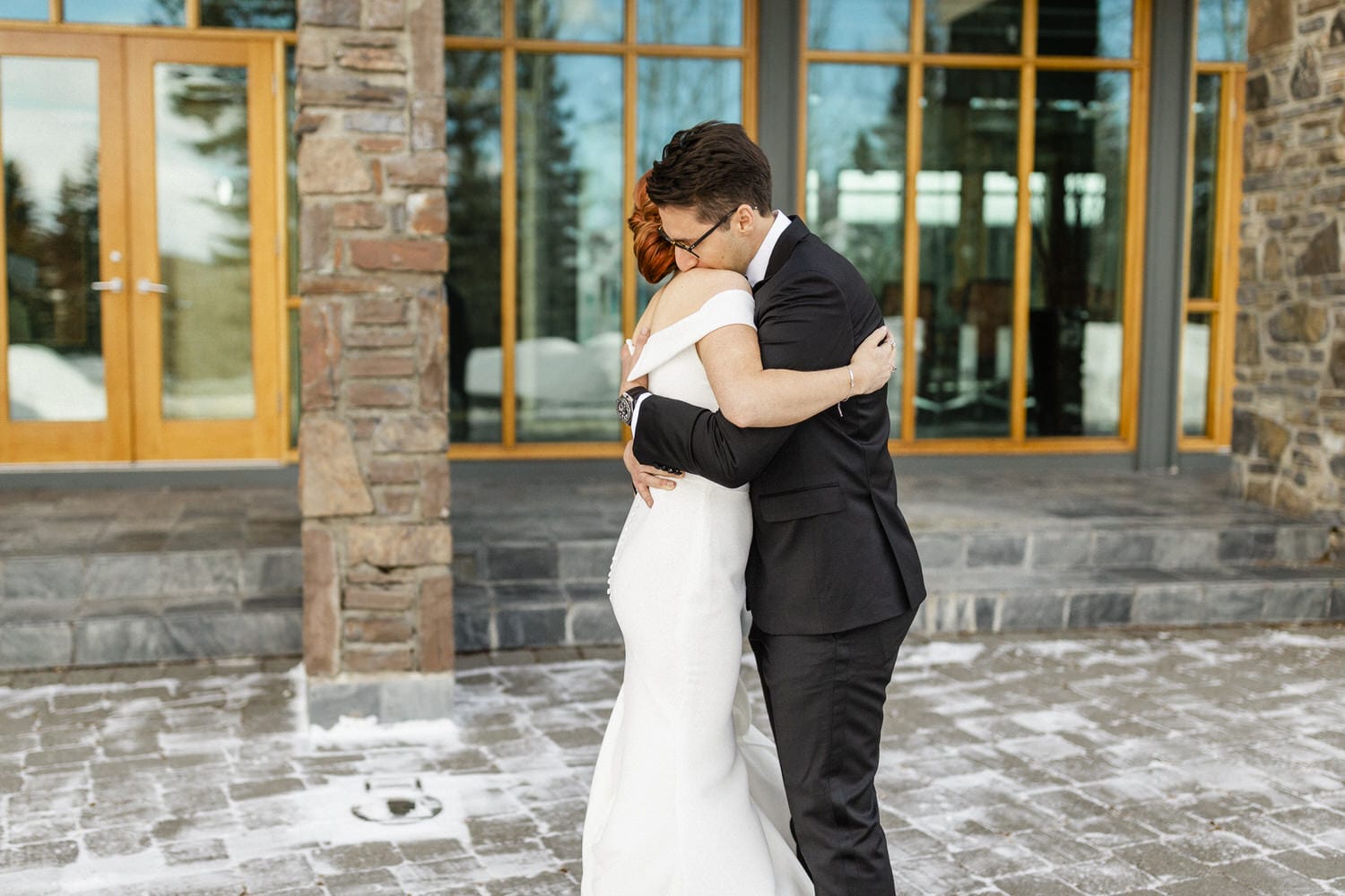 A couple sharing a warm embrace outside a modern stone and wood building, surrounded by winter scenery and snow.