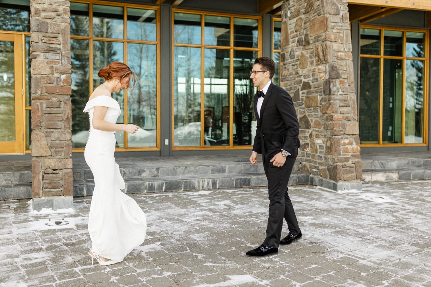 A couple moments before their wedding ceremony, with the bride in a stunning white dress and the groom in a classic black tuxedo, standing outside a modern stone building.