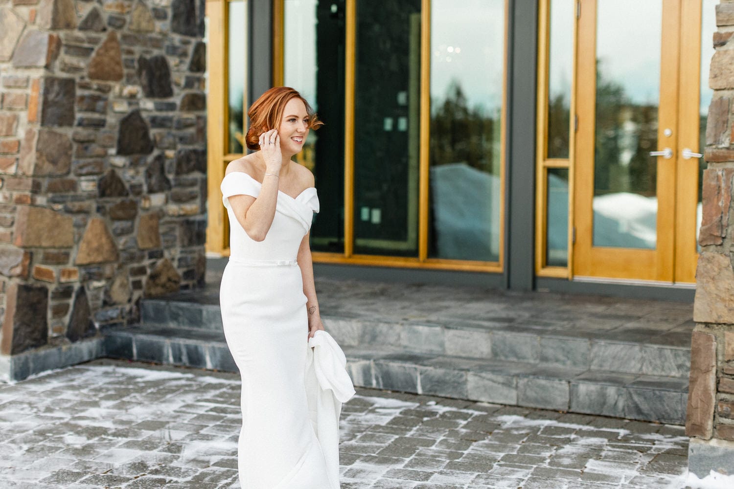 A joyful bride in a fitted white gown with off-the-shoulder sleeves, standing outside a modern stone building, holding her veil and smiling while talking on the phone.