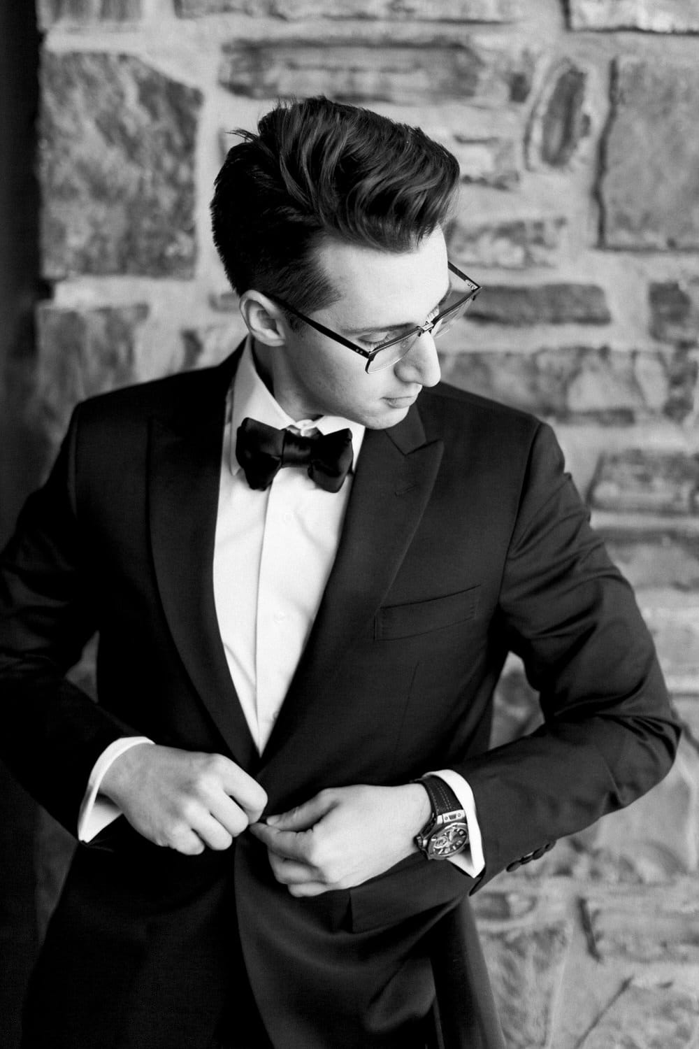 A young man adjusts his bow tie and buttons his tuxedo jacket, showcasing a sophisticated look against a stone wall in a black and white photograph.