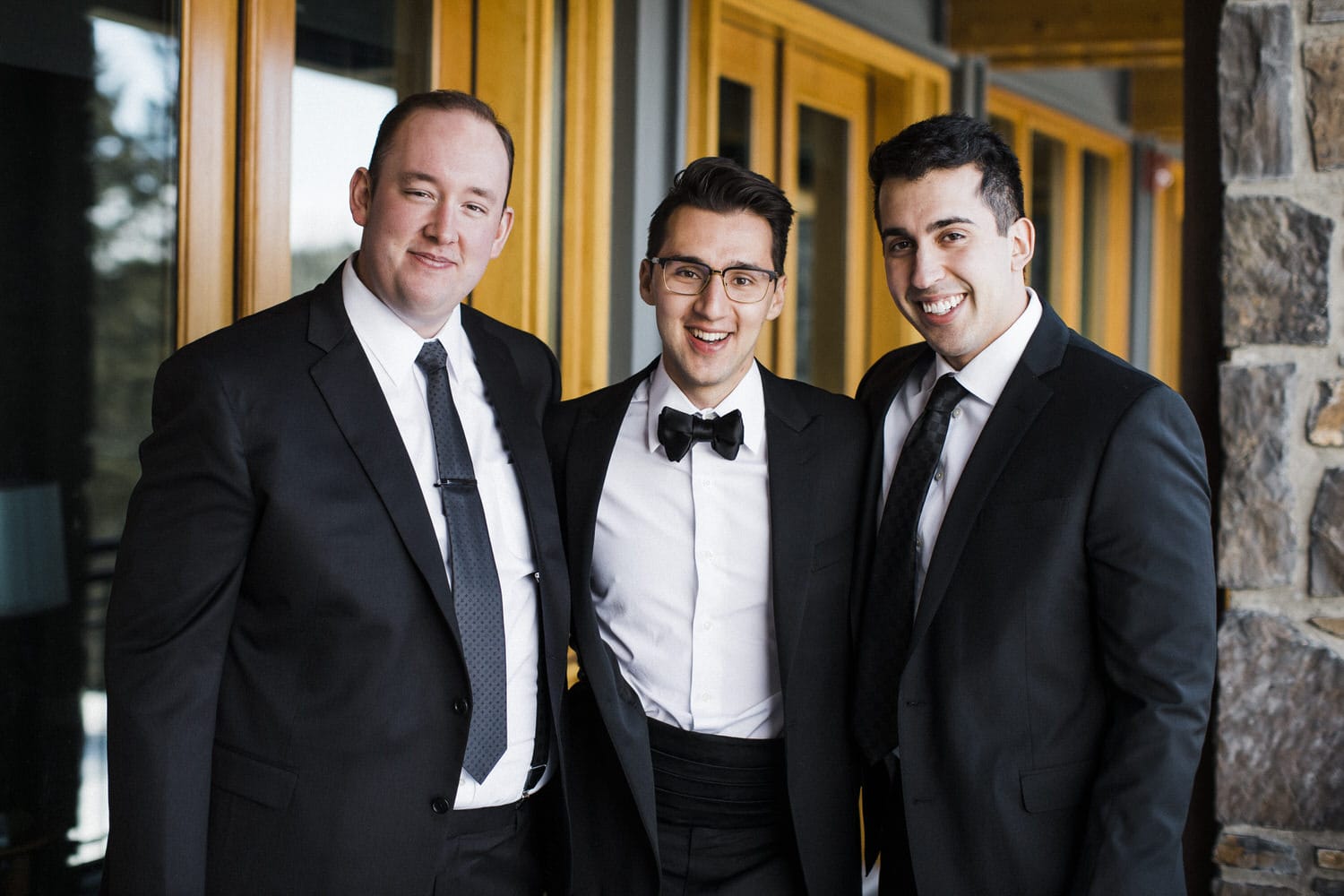 Three men in tuxedos pose happily together against a backdrop of wooden doors and a stone wall.