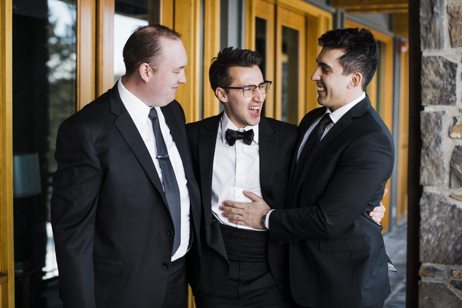 Three men in suits share a joyful moment, smiling and embracing each other against a backdrop of wooden doors.